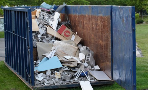 A large, open-top industrial container positioned outdoors on a grassy area, filled with a mixture of construction debris, including broken plywood, plasterboard, cement blocks, and various other building materials. The container's sides are painted dark blue, with visible signs of rust and wear on the steel surface. Inside, the materials are piled haphazardly, with some items leaning against the sides and others scattered across the base. The debris appears weathered, with rough textures and dull colours, predominantly light browns, greys, and off-white shades, indicating discarded building rubbish. In the background, there is a well-maintained green lawn and shrubbery, suggesting a suburban or semi-rural setting typically serviced by local waste removal companies such as Commercial Waste Removal Downham. The scene reflects a typical rubbish removal scenario, where a professional service would be needed to clear construction waste from a site in the Downham area, aligning with the company's expertise in managing bulky waste disposal efficiently and responsibly.