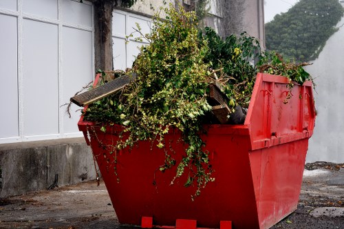 A large red skip bin filled with various types of garden waste, including green leafy branches, small trees, and plant debris, positioned on a paved surface near a white panelled wall with a concrete base. The waste appears to be freshly collected, with some branches and foliage spilling over the edges of the bin. The environment suggests an outdoor urban or suburban setting, likely associated with domestic or commercial waste removal services. The scene is well-lit, highlighting the textures of the green foliage, the smooth, bright red finish of the skip, and the surrounding paved ground, reflecting a typical rubbish collection or clearance activity in the Downham area. The image is relevant to Commercial Waste Removal Downham, illustrating services related to garden or green waste disposal within the local context.